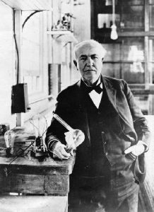 A white-haired man in coat and tie holding a lightbulb in a lab.