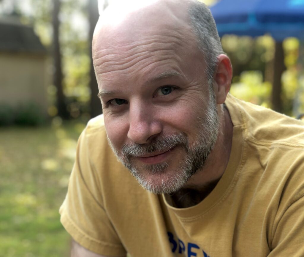 Casual head shot for Charlie Bennett, a middle-aged man with trimmed beard and bald head in a sunlit yard.